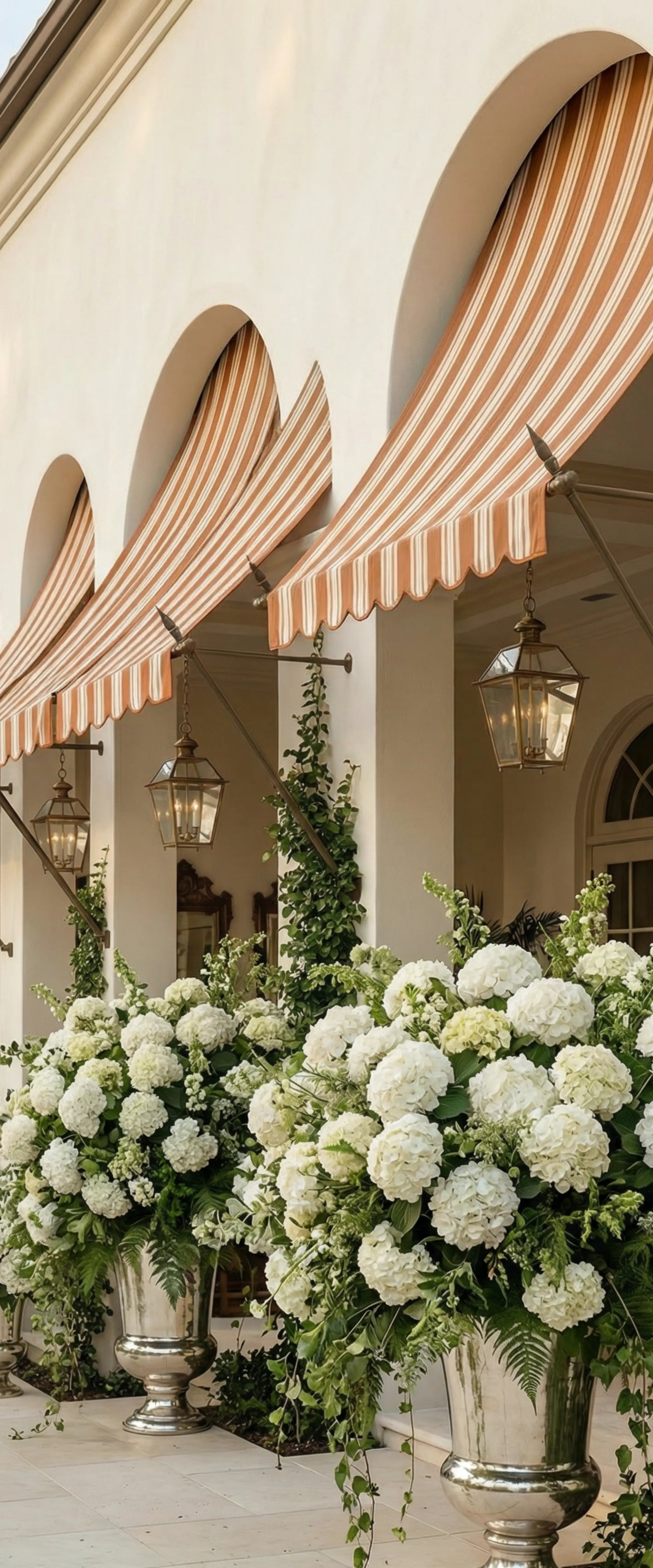 Banner décoratif Dolce Arco - Façade italienne avec arcades en pierre beige, stores rayés terracotta et blanc, lanternes laiton, grandes urnes argentées débordant d'hortensias blancs et lierre, ambiance palazzo luxueux