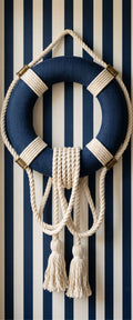 Banner décoratif L'escale - Bouée de sauvetage bleu marine avec cordages naturels et pompons macramé sur mur en papier peint rayé bleu marine et blanc, ambiance nautique épurée