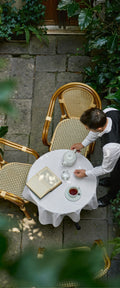 Banner décoratif Café discret - full - Vue aérienne terrasse parisienne avec fauteuils rotin cannage, table ronde nappe blanche, thé, livre, pavés anciens, plantes, ambiance café chic